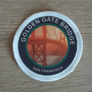 Könnte beinhalten: Ein weißer, runder Aufnäher mit schwarzem Rand und einem roten und orangefarbenen Bild der Golden Gate Bridge. Der Text "Golden Gate Bridge" ist in Schwarz um das Bild herum geschrieben und "San Francisco" ist in Schwarz unter dem Bild geschrieben.
