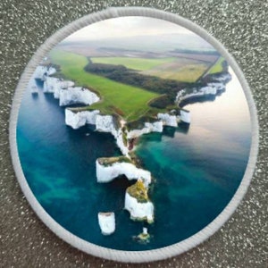 May include: Aerial view of a white rock formation jutting out into a turquoise blue ocean. The rock formation is surrounded by green grass and trees.