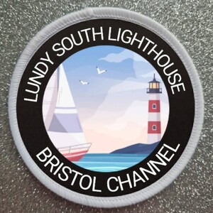 以下が含まれることがあります： 縁に「Lundy South Lighthouse Bristol Channel」という文字が書かれた白黒の円形のワッペン。ワッペンの中央には、青い空を背景に、灯台、帆船、カモメが描かれたカラフルなイラストが描かれている。