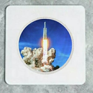 Könnte beinhalten: Eine weiße Rakete mit einer leuchtend orangefarbenen Flamme startet in einen blauen Himmel mit weißen Wolken.