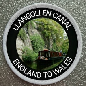 May include: Round patch with the words "LLANGOLLEN CANAL ENGLAND TO WALES" in white. The patch shows a canal boat on a waterway, with green trees and a rocky cliff in the background.