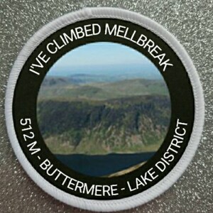 May include: White round patch with black text and a photo of a mountain range and lake. The text reads "I've Climbed Mellbreak 512 M - Buttermere - Lake District".