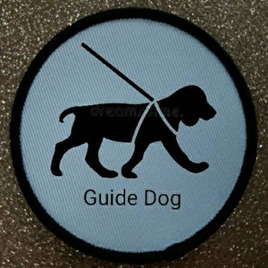 Puede incluir: Un parche redondo azul y negro con una silueta negra de un perro guía con arnés y correa. El texto "Guide Dog" está impreso en negro debajo del perro.