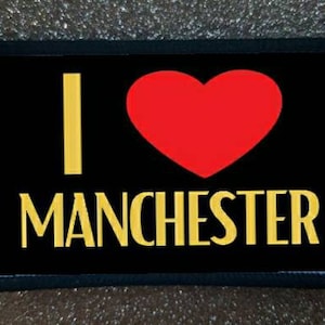 May include: A black rectangular patch with the words "I MANCHESTER" in gold lettering and a red heart symbol. The patch has a black border and is set against a dark background.