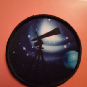 Może przedstawiać: Okrągła naszywka w kolorze czarnym i niebieskim z sylwetką teleskopu na statywie na tle nocnego, gwieździstego nieba.
