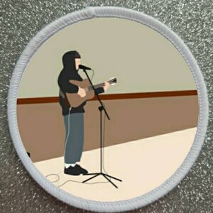 May include: A white patch with a brown and beige background. A person wearing a black hooded sweatshirt is standing on a stage holding a brown acoustic guitar and singing into a microphone. The person is standing in front of a microphone stand.