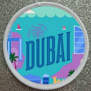 Op de afbeelding: Een ronde witte patch met een blauwe rand en het woord "DUBAI" in blauwe letters. De achtergrond is turquoiseblauw met een roze en paarse stadsgezicht en palmbomen.