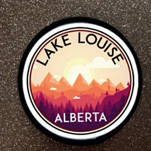 Könnte beinhalten: Ein runder Aufkleber mit weißem Hintergrund und schwarzem Rand. Der Aufkleber zeigt eine stilisierte Illustration von Bergen, einer Sonne und Bäumen in Orange-, Rot- und Lilatönen. Der Text "LAKE LOUISE ALBERTA" ist in Schwarz auf dem Aufkleber gedruckt.