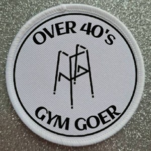 Puede incluir: Parche redondo blanco con texto negro y contorno negro. El texto dice "Over 40's Gym Goer" y presenta una silueta negra de un andador.