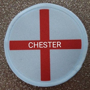 Op de afbeelding: Een ronde patch met een lichtblauwe achtergrond en een rood kruisontwerp. Het woord "CHESTER" is in het wit over het midden van het kruis gedrukt. De patch heeft een lichtblauwe rand.