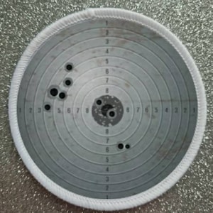 Può includere: Un bersaglio circolare bianco con cerchi concentrici e numeri da 1 a 8. Il bersaglio ha diversi fori di proiettile al centro.