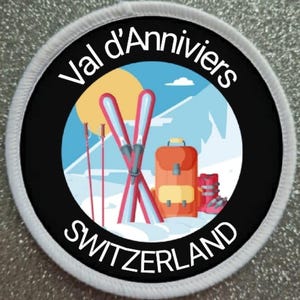 Può includere: Un cerotto circolare bianco e nero con il testo "Val d'Anniviers SWITZERLAND" attorno al bordo. Il centro del cerotto presenta un'illustrazione di cartone animato di una scena di montagna con sci, bastoncini da sci, uno zaino e scarponi da sci.
