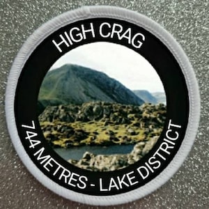 May include: White round patch with black border and white text. The text reads "HIGH CRAG 744 METRES - LAKE DISTRICT". A photo of a mountain range is inside the circle.