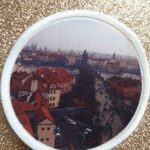 May include: A round white patch with a printed image of a cityscape. The image shows a view of a city with red-roofed buildings and a river in the foreground. The background shows a hazy sky with a few buildings in the distance.