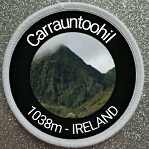 May include: White round patch with a black circle in the center. The black circle has white text that reads "Carrauntoohil 1038m - IRELAND" and a photo of a mountain peak in the center.
