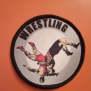Könnte beinhalten: Ein schwarz-weißes Wrestling-Patch mit dem Wort "WRESTLING" in einem Kreis um zwei Ringer in einem Griff. Die Ringer tragen rote und blaue Wrestling-Anzüge.