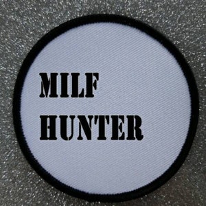 Op de afbeelding: Ronde witte patch met zwarte geborduurde tekst die "MILF HUNTER" luidt.