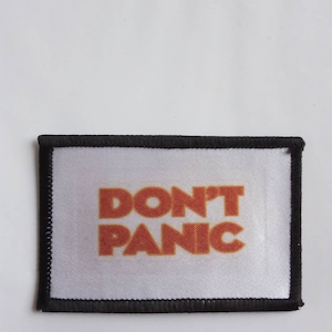 Peut inclure: Un patch en tissu blanc avec des coutures noires autour des bords. Le patch a les mots "DON'T PANIC" imprimés en orange.