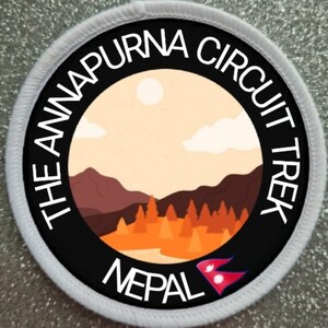 Puede incluir: Un parche redondo blanco y negro con el texto "The Annapurna Circuit Trek Nepal" alrededor del borde. Dentro del círculo hay una ilustración colorida de una cordillera con un río, árboles y un sol en el cielo.