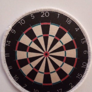 Puede incluir: Un tablero de dardos circular blanco con anillos negros y rojos y un anillo exterior verde. Los números del 1 al 20 están impresos alrededor del borde del tablero.