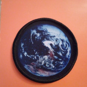 Könnte beinhalten: Ein runder, schwarz umrandeter Patch mit einem gedruckten Bild der Erde in Blau- und Weißtönen.