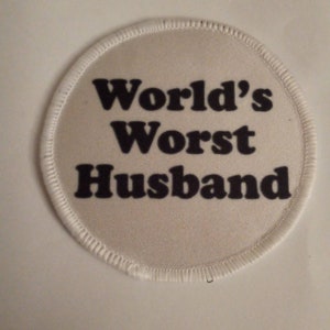 May include: A circular white fabric patch with a stitched border. The text "World's Worst Husband" is printed in bold black capital and lowercase letters across the center of the patch, occupying most of the circular surface area.
