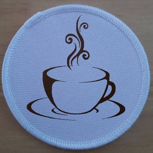 May include: A white round fabric patch with a brown outline and a brown silhouette of a coffee cup with steam rising from it.