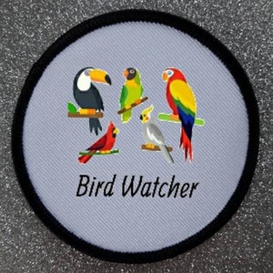 Könnte beinhalten: Ein runder, weißer Patch mit schwarzem Rand zeigt eine bunte Illustration von fünf Vögeln. Die Vögel umfassen einen Tukan, einen Kardinal, einen grünen Papagei, einen gelben und roten Papagei und einen Nymphensittich. Der Text "Bird Watcher" ist in Schwarz unter den Vögeln gedruckt.