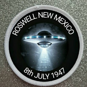 May include: A white round patch with black text and a black circle in the center. The text reads "Roswell New Mexico 8th July 1947". The black circle contains a black and white image of a flying saucer.