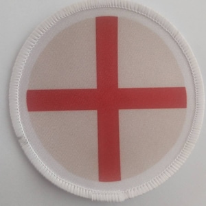 May include: A white round patch with a red cross in the centre. The patch has a white stitched border.