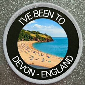 May include: A white round patch with black text that reads "I've Been To Devon - England". The patch features a photo of a beach with blue water and a green cliff in the background.