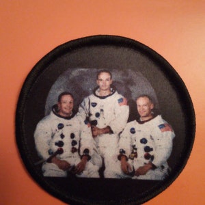 Könnte beinhalten: Ein Schwarzweißfoto von drei Astronauten in ihren Raumanzügen. Die Astronauten stehen vor einem Hintergrund der Mondoberfläche. Das Foto befindet sich auf einem runden, schwarzen Patch mit schwarzem Rand.