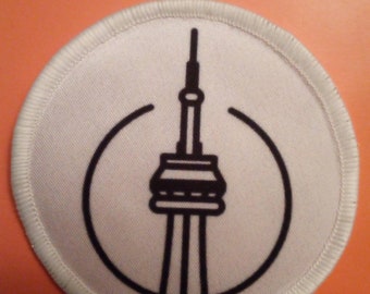 May include: White round fabric patch with a black outline of the CN Tower. The patch has a white background and a black outline of the CN Tower.