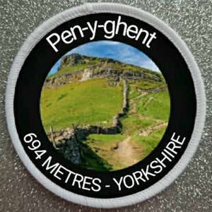Könnte beinhalten: Ein runder weißer und schwarzer Aufnäher mit einem Foto eines grünen Hügels und einer Steinmauer. Der Text "Pen-y-ghent 694 Meter - Yorkshire" ist auf dem Aufnäher aufgedruckt.