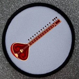 Könnte beinhalten: Ein runder Aufnäher mit einer Sitar, einem Saiteninstrument. Die Sitar ist in Rot- und Brauntönen vor einem hellblauen Hintergrund dargestellt. Der Aufnäher hat einen schwarzen Rand.
