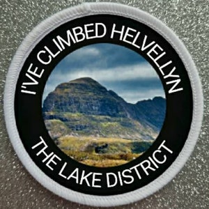 May include: A white round patch with black text and a photo of a mountain range. The text reads "I've Climbed Helvellyn The Lake District".