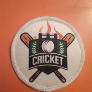 Könnte beinhalten: Runder weißer Patch mit einem Cricket-Logo. Das Logo zeigt zwei gekreuzte Schläger, einen Ball und einen grün-schwarzen Schild mit dem Wort "CRICKET" in weißen Buchstaben. Der Schild ist mit einer roten und orangefarbenen Flamme gekrönt.