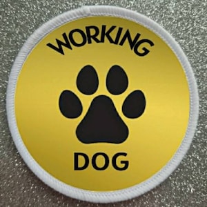 Könnte beinhalten: Ein weißer runder Patch mit gelbem Hintergrund und schwarzem Text. Der Text lautet "WORKING DOG" mit einem schwarzen Pfotenabdruck in der Mitte.