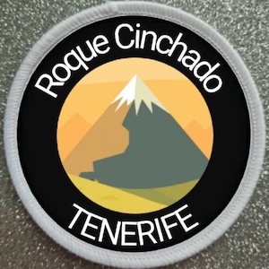 Puede incluir: Un parche circular blanco con texto negro y un círculo negro en el centro. El texto dice "Roque Cinchado TENERIFE". El círculo negro contiene una ilustración estilizada de una montaña con un pico blanco sobre un fondo amarillo.