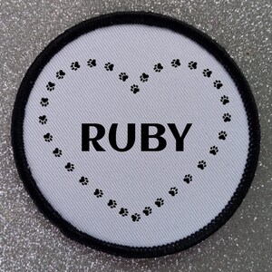 May include: White round patch with a black border. The patch has a heart shape made of paw prints with the name "RUBY" in black letters inside the heart.