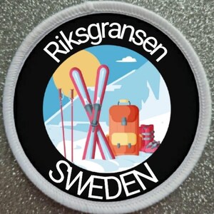 Peut inclure: Un écusson circulaire blanc avec du texte noir qui dit "Riksgransen SWEDEN". Le centre de l'écusson présente une illustration colorée d'une scène de montagne enneigée avec deux paires de skis, des bâtons de ski et un sac à dos rouge.