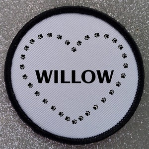 May include: White round fabric patch with a black border. The patch has a heart shape made of paw prints with the word "WILLOW" in the center.