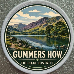 May include: A circular embroidered patch featuring a landscape illustration of Gummers How in the Lake District. The scene depicts a calm lake, green hills, and mountains under a blue sky with clouds. The text GUMMERS HOW THE LAKE DISTRICT is printed.