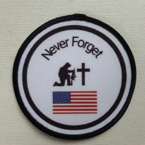 May include: A round black and white patch with the words "Never Forget" in a circle around a silhouette of a person kneeling with a cross and an American flag.
