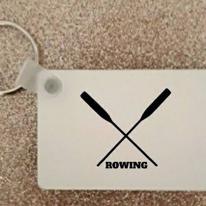 May include: White keychain with a black silhouette of two crossed oars and the word "ROWING" underneath.