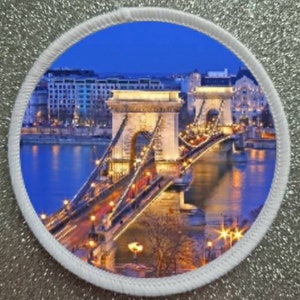 May include: A night view of the Chain Bridge in Budapest, Hungary. The bridge is lit up with lights, and the city skyline is visible in the background.