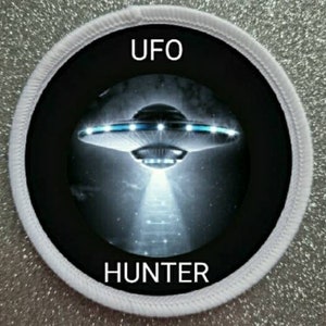 Op de afbeelding: Ronde witte patch met zwarte rand en een afbeelding van een vliegende schotel met de tekst "UFO HUNTER" in wit.