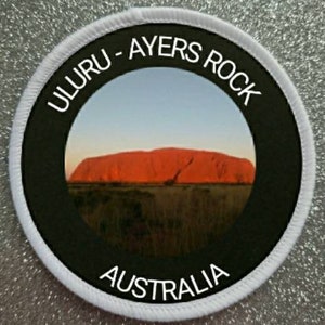 Può includere: Una toppa rotonda bianca e nera con il testo "Uluru - Ayers Rock Australia" attorno a una foto di Uluru, una grande formazione rocciosa rossa in Australia.