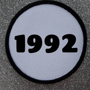 Könnte beinhalten: Ein runder Aufnäher mit hellgrauem Hintergrund und schwarzem Rand. Die Zahl "1992" ist in fetter schwarzer Schrift mittig auf dem Aufnäher aufgedruckt. Der Aufnäher ist wahrscheinlich ein bestickter oder bedruckter Artikel.