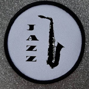 Pode incluir: Um remendo circular branco com uma borda preta. O remendo apresenta uma silhueta preta de um saxofone com a palavra "JAZZ" impressa verticalmente em preto.